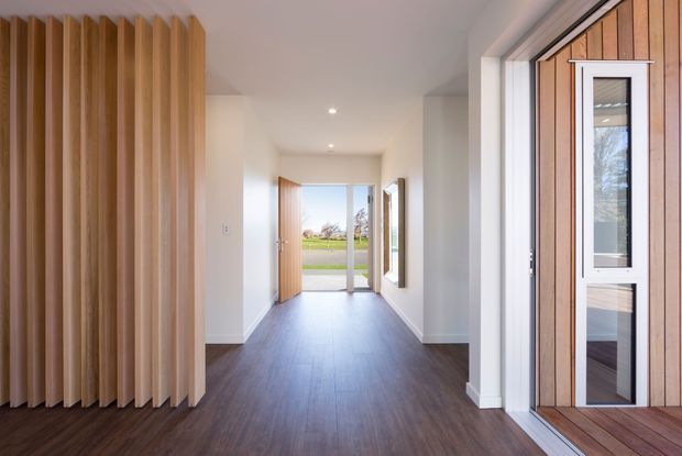Bright contemporary entry hall with dark timber flooring, white walls and recessed ceiling lights. A full-height vertical timber slat feature screens the left side, while a glazed timber-framed entrance door and adjacent glazing open toward the exterior. The hallway extends to a rear glazed door with views to a lawn and road beyond.