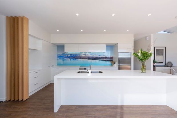 Bright contemporary kitchen with a large white island and integrated sink, white full-height cabinetry, dark timber-look flooring, stainless-steel refrigerator, and a blue scenic feature splashback depicting a mountain-and-water landscape. Vertical timber slats form a feature screen at the left, while recessed ceiling lights provide even illumination.