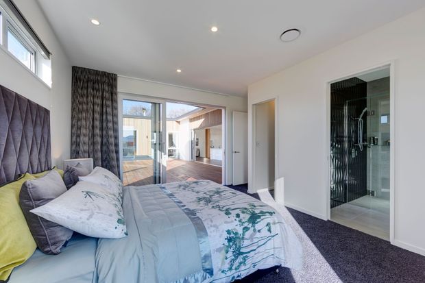 Bright contemporary bedroom with a large upholstered quilted headboard, layered blue, grey and lime bedding, dark patterned curtains, and dark carpet. Glazed doors open to a timber deck and courtyard, while a tiled bathroom or ensuite is visible through the opening on the right. The room has recessed ceiling lights, white walls, and high-level windows.
