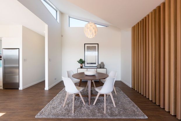 Bright contemporary dining area with a round timber dining table and white molded chairs on a textured rug. The room features dark timber flooring, white walls, a sculptural pendant light, skylights, minimalist artwork and decor, and a prominent full-height curved timber slat feature wall on the right. A stainless-steel refrigerator and adjacent hallway are visible in the background.