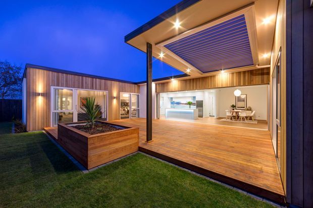 Contemporary single-storey home photographed at dusk, featuring a large timber deck and covered outdoor entertaining area with a slatted ceiling and recessed lighting. Full-height glazed doors open to a bright open-plan interior with a visible kitchen and dining area. Timber-clad walls, a raised garden planter, and integrated indoor-outdoor living create a warm modern setting.