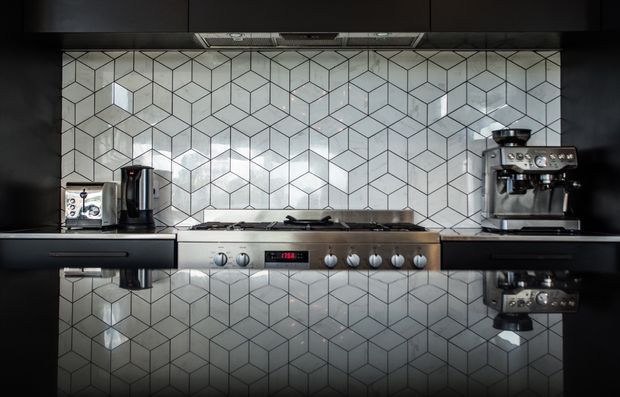 A dark, modern kitchen benchtop composition featuring a stainless-steel gas cooktop and oven, black cabinetry, a reflective black benchtop, and a geometric white-and-grey hexagonal tile splashback. A kettle and espresso machine sit on either side of the cooktop, with strong reflections visible on the benchtop.