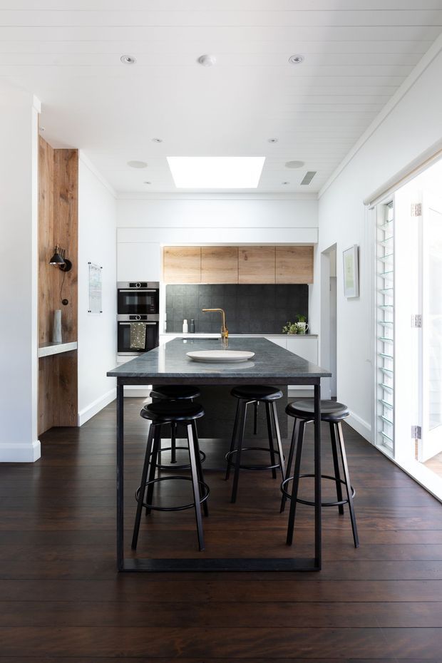 Bright contemporary kitchen with a large dark stone island and integrated dining-height bench, surrounded by black metal stools. The room features dark timber flooring, white walls and cabinetry, warm timber overhead cupboards, a dark tiled splashback, recessed lighting, and abundant daylight through louvred glazing and glazed doors.