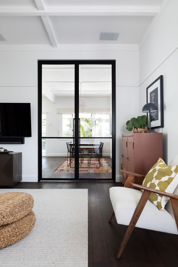 Bright contemporary living room with dark timber flooring, white walls and exposed ceiling beams. The room features a wall-mounted television, pale area rug, woven coffee table, mid-century-style upholstered lounge chair, framed artwork, and a muted rose built-in cabinet. Black-framed glazed double doors open to a dining area with a table, chairs, and patterned rug; greenery is visible beyond the glazing.