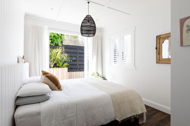 Bright, compact bedroom with a white upholstered or panelled bed, layered white bedding, a beige throw, and a woven black pendant light. White plantation shutters and sheer curtains provide abundant daylight, while a timber-framed mirror, dark timber flooring, and a small indoor planter add warmth. The room has a relaxed, coastal-inspired contemporary feel.