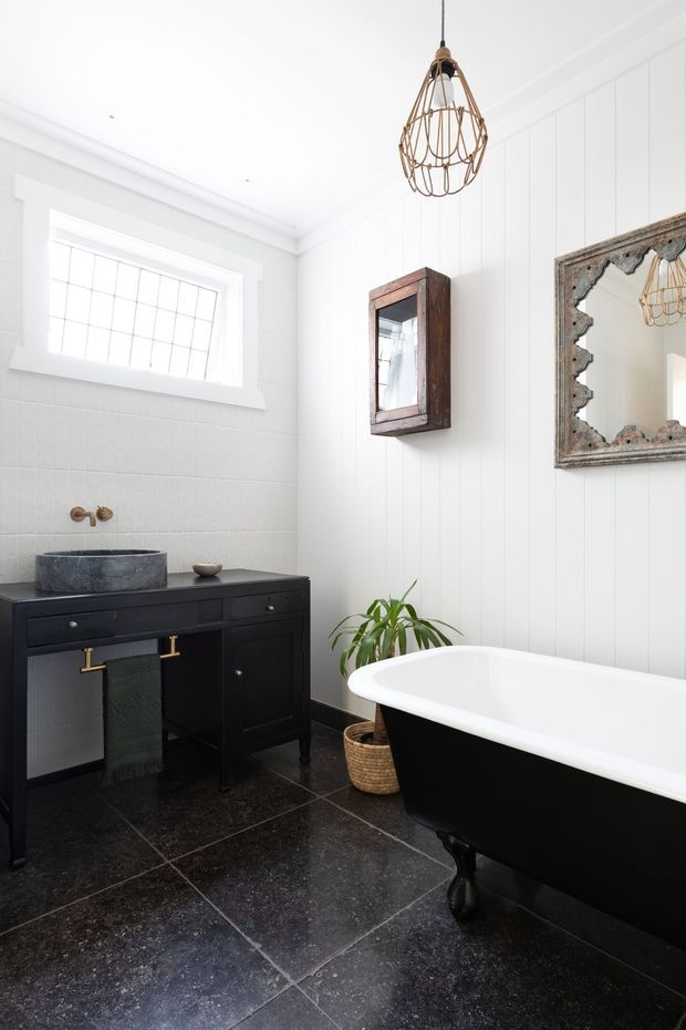 Bright, refined bathroom featuring a freestanding black-and-white clawfoot bathtub, black stone-look floor tiles, a dark timber vanity with a stone vessel basin, white vertical wall panelling, a leaded-glass window, vintage mirrors, and an ornate wire pendant light.