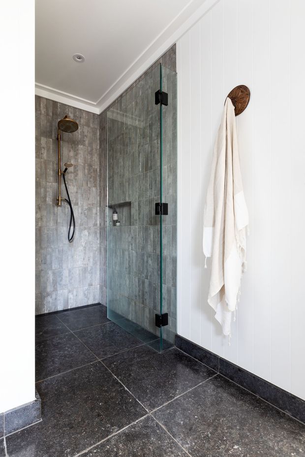 A contemporary walk-in shower bathroom with a frameless glass screen, wall-mounted rainfall and handheld shower, dark large-format floor tiles, grey vertically textured wall tiles, and white vertical wall panelling. A timber towel hook and white towel add warmth to the otherwise monochromatic space.