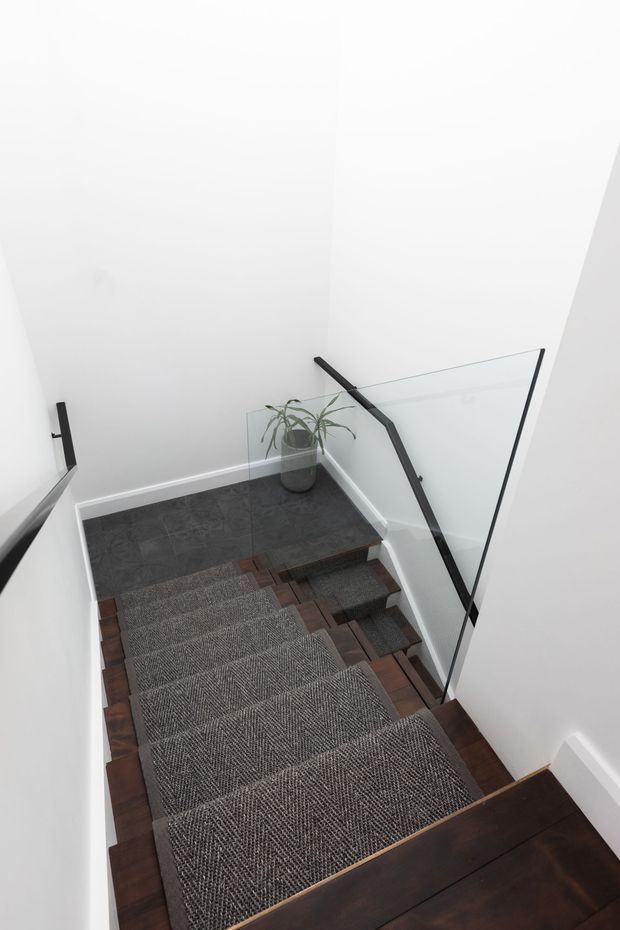 A modern residential stairway viewed from above, with dark timber treads, grey patterned stair runners, white walls, dark tiled flooring at the landing, and a frameless glass balustrade supported by black metal fittings. A potted plant adds a small natural accent.