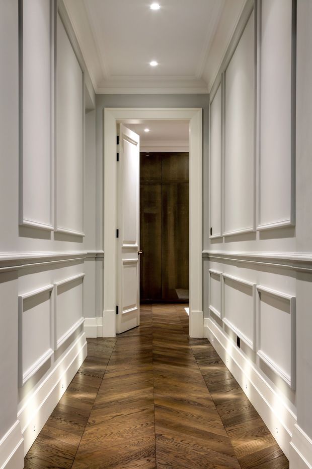 A narrow, refined residential hallway with white decorative wall panelling, raised mouldings, concealed cabinetry or panelled doors, recessed ceiling lights, and a warm dark timber herringbone floor. The corridor leads through an open doorway to a darker timber-lined space, creating strong depth and contrast.