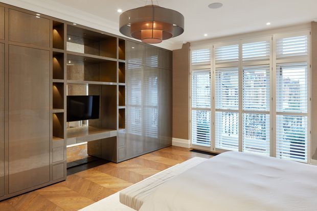 A refined bedroom with custom built-in timber cabinetry, open display shelving, integrated television, and a wardrobe wall. The room features warm herringbone timber flooring, white walls and ceiling, plantation shutters over large windows, and a sculptural circular pendant light. A bed occupies the foreground, creating a warm, polished and contemporary-classic atmosphere.