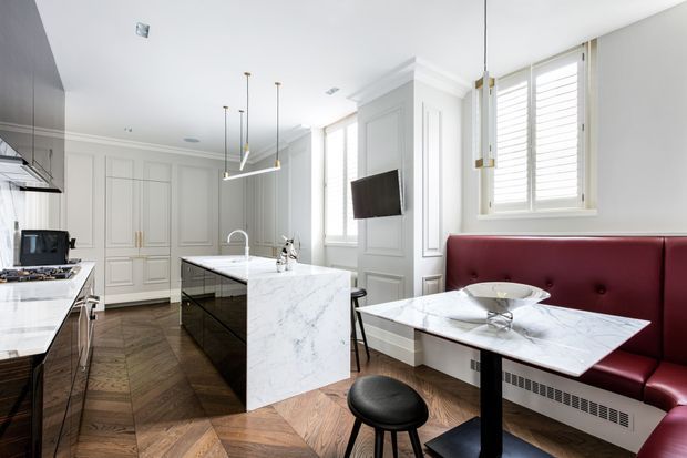 Bright, refined kitchen and dining nook with white paneled cabinetry, a large marble-look island, dark chevron timber flooring, and a built-in burgundy leather banquette beside marble-topped dining tables. Shuttered windows, decorative wall mouldings, and pendant lighting add a polished classic feel.