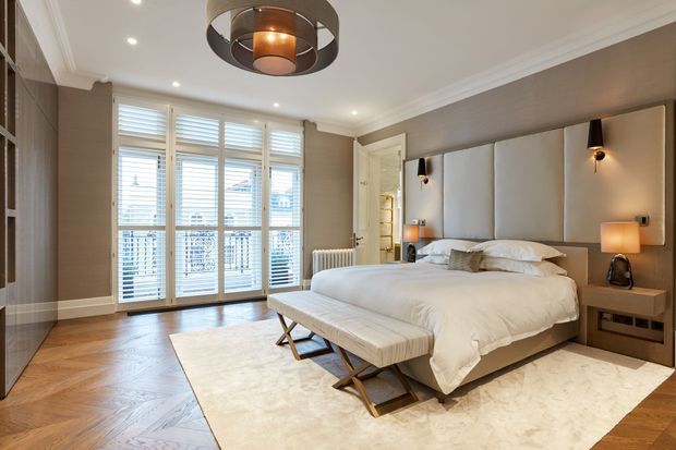 A refined contemporary bedroom with a large upholstered bed, padded full-width headboard, matching bedside tables, and a bench at the foot of the bed. The room features warm herringbone timber flooring, a large pale rug, taupe textured walls, white decorative ceiling moulding, plantation shutters across full-height glazed doors or windows, integrated wardrobe cabinetry, and layered pendant and wall lighting.
