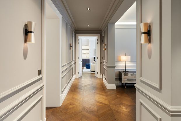 Elegant residential hallway with warm herringbone timber flooring, pale grey decorative wall panelling and mouldings, integrated wall sconces, and views into a refined sitting area and adjoining room. The corridor has a balanced, classic interior with a contemporary lighting scheme.