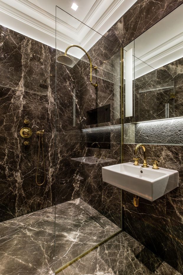 A compact, luxurious bathroom with dark brown veined marble walls and flooring, a frameless glass walk-in shower, wall-mounted white basin, large mirror, concealed black fixtures, and polished brass shower and tapware. Decorative ceiling mouldings and integrated lighting add a refined, dramatic character.