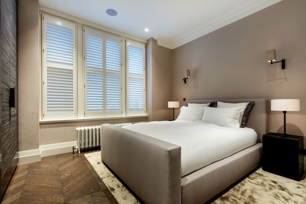 A refined bedroom with a large upholstered bed dressed in white linens, dark bedside tables, matching table lamps, and wall-mounted sconces. The room features warm taupe walls, white decorative ceiling moulding, plantation shutters across the windows, a vintage-style radiator, herringbone timber flooring, and a pale patterned area rug. The overall look is calm, polished, and contemporary-classic.