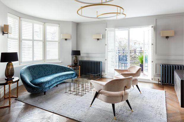 A refined living room with ornate wall mouldings, tall shuttered windows, French doors opening to a balcony, dark cast-iron radiators, herringbone timber flooring, and a large pale area rug. The room is furnished with a curved teal velvet sofa, two cream upholstered armchairs, glass-and-metal coffee tables, sculptural lamps, and a striking circular brass pendant light.