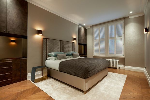 A sophisticated bedroom with a central upholstered bed, textured grey-brown bedding, integrated dark timber cabinetry and bedside lighting. The room features herringbone timber flooring, a pale area rug, a textured neutral wall finish, white decorative cornices, plantation shutters, and a traditional radiator beneath the window.