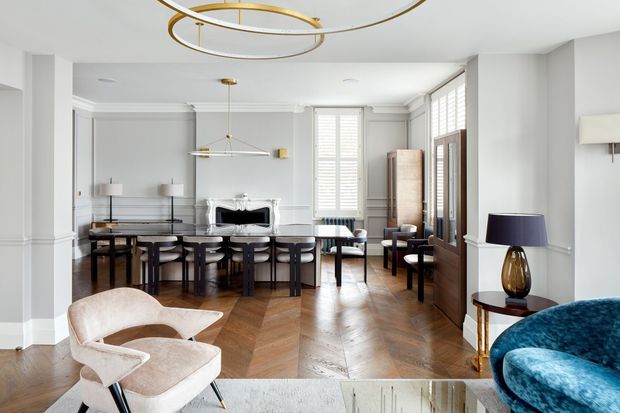 Bright, refined open-plan interior combining a formal dining area with an adjoining living space. The room features white decorative wall panelling and ornate cornices, herringbone timber flooring, tall shuttered windows, dark dining furniture, a marble fireplace, and brass-accented contemporary lighting. A blue velvet occasional chair and neutral sculptural armchair add contrasting colour and texture.