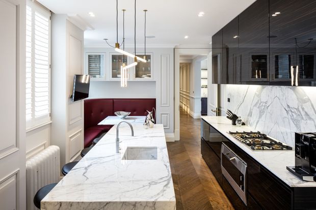 Refined contemporary kitchen and dining nook with a large white marble island, integrated sink and seating, dark timber cabinetry, marble splashback, brass pendant lights, and a burgundy upholstered banquette. White paneled walls, plantation shutters, herringbone timber flooring, and a hallway create a polished, detailed interior.