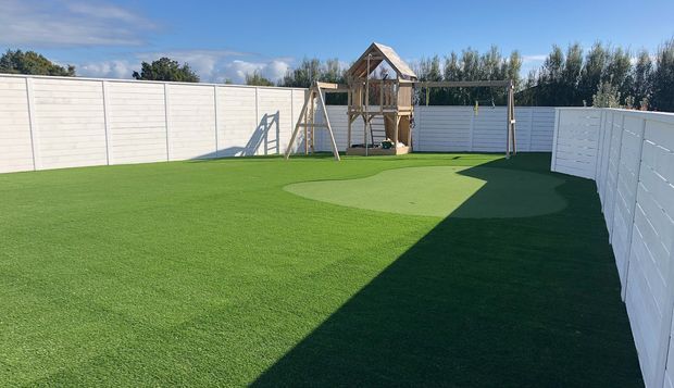 A spacious enclosed backyard with a large artificial-turf lawn, white horizontal-panel perimeter fencing, and a timber children's play structure with a small pitched roof and swing set. Strong sunlight creates a broad shadow across the lawn, with trees visible beyond the fence.