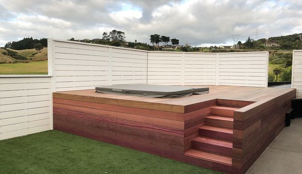 Elevated outdoor deck area with a raised, built-in timber platform and integrated steps, enclosed by white horizontal privacy fencing. A low, dark-covered rectangular feature sits on the platform, with artificial lawn in the foreground and a rural landscape beyond.
