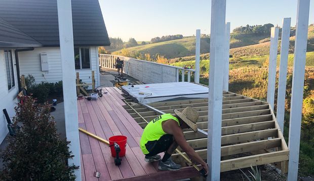 A residential outdoor deck under construction beside a light-clad house. Workers are assembling a timber-framed deck extension with exposed joists and tall white posts, while completed reddish timber decking and a partially installed hot tub or spa area are visible. Rolling rural farmland is seen beyond the deck.