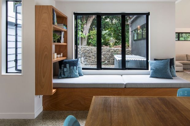 Bright contemporary interior featuring a built-in window seat with white cushions and blue pillows, integrated warm timber shelving and cabinetry, black-framed windows, and a timber dining table in the foreground. A landscaped stone retaining wall and greenery are visible outside, while part of an adjoining seating area appears at right.