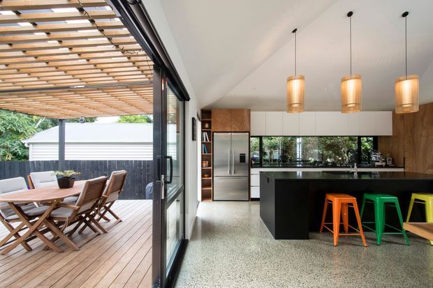 Contemporary open-plan kitchen and dining area opening onto a timber deck through large sliding glass doors. The kitchen features a dark island with colourful stools, white upper cabinetry, timber wall cabinetry, stainless-steel appliances, a terrazzo-like floor, and pendant lighting. A timber pergola shades the outdoor dining area.