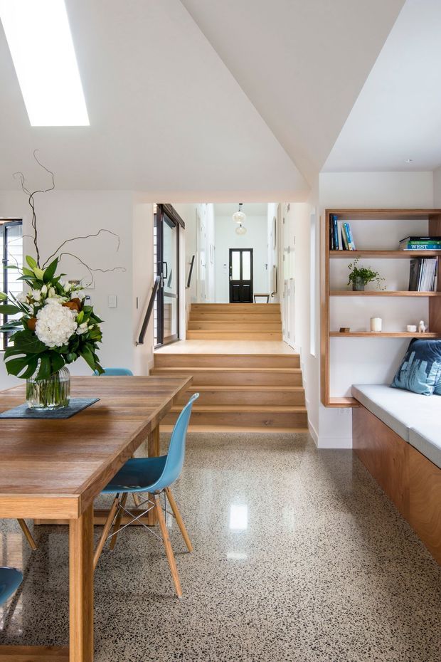 Bright open-plan interior featuring a timber dining table and blue chairs in the foreground, polished speckled terrazzo flooring, a timber stair leading to a hallway, and built-in timber shelving with a window seat on the right. A large skylight and clerestory-style glazing provide abundant natural light.