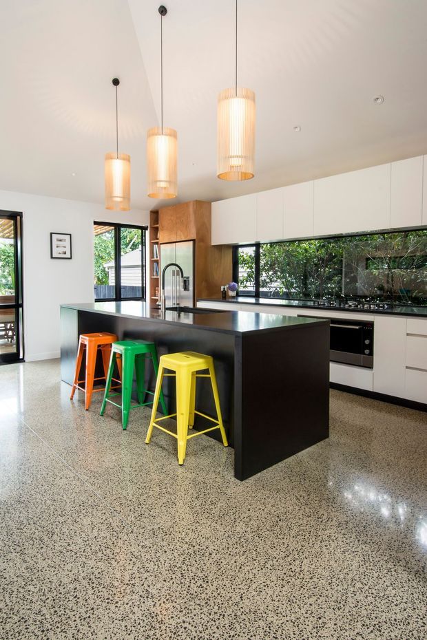 Bright contemporary kitchen with a large black island and waterfall-style benchtop, colourful metal bar stools, white flat-panel cabinetry, timber cabinetry, integrated appliances, and a wide horizontal window overlooking greenery. Three pendant lights hang above the island, and the polished speckled concrete or terrazzo-like floor extends through the open space.