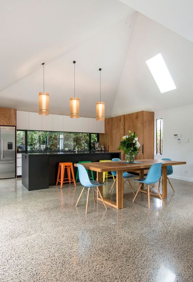 A bright, open-plan kitchen and dining area with a high sloping ceiling. The kitchen features matte black cabinetry, white upper cabinets, a timber-panelled wall, dark benchtops, and extensive garden-facing glazing. Three suspended warm pendant lights hang over the island, while a large timber dining table is surrounded by colourful modern chairs. The space has a polished speckled terrazzo or polished concrete floor, a skylight, and a clean contemporary feel.