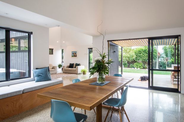 Bright open-plan dining and living area with a large timber dining table, blue dining chairs, built-in window seat, polished terrazzo-style flooring, and expansive black-framed glazing opening to a landscaped outdoor area. White walls and ceilings create an airy contemporary interior, with a covered pergola visible outside.