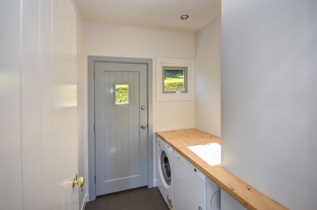 Compact laundry and entry area with a grey paneled exterior door, small window, white built-in cabinetry, front-loading washing machine, and a warm timber benchtop. The space has white walls and cabinetry, dark floor tiles, and recessed ceiling lighting.