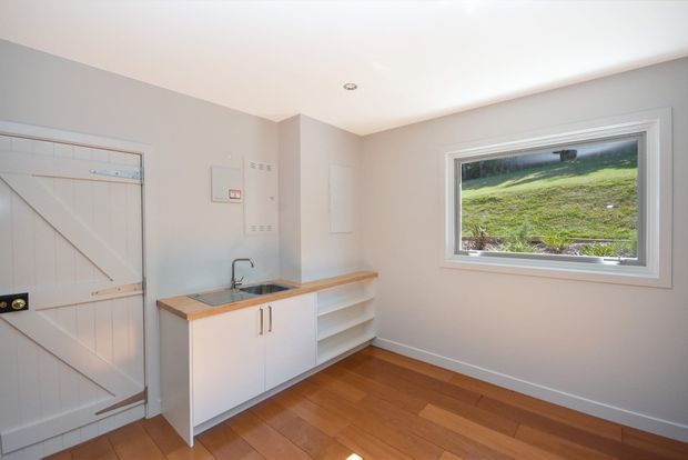 Bright compact utility room with white built-in cabinetry, open shelving, a timber benchtop and integrated stainless-steel sink. The room has warm timber flooring, pale walls, a white barn-style door, and a large window overlooking a grassy landscape.
