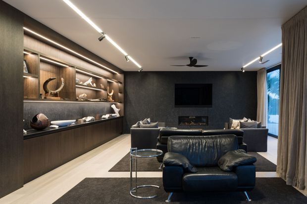 A sophisticated contemporary living room with a dark timber built-in display wall and cabinetry, integrated shelving, a large wall-mounted television, and a linear fireplace set into a dark textured feature wall. The room includes deep navy leather seating, a sofa arrangement, a glass-and-metal side table, recessed display lighting, track lighting, full-height curtains, and pale timber flooring. The overall atmosphere is refined, dark, and cinematic.