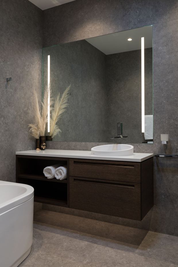 A sophisticated modern bathroom featuring dark textured stone-look wall and floor finishes, a floating dark timber vanity with integrated open shelving, a white engineered-stone benchtop, vessel basin, wall-mounted tapware, and a large illuminated mirror. A freestanding white toilet and subtle decorative dried grasses complete the space.