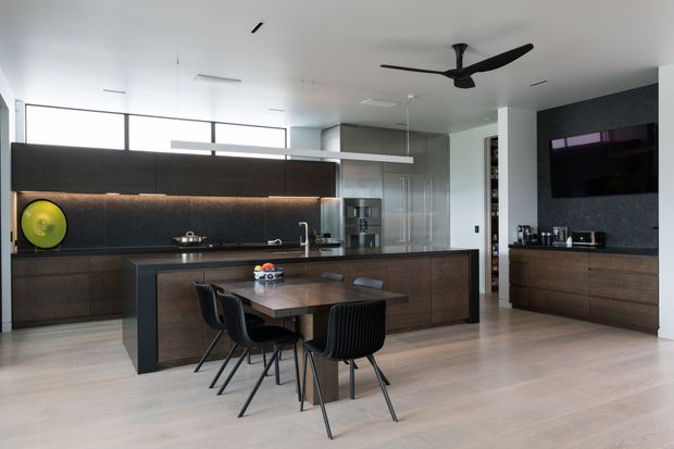 Contemporary open-plan kitchen and dining area with extensive dark timber cabinetry, a large central island, integrated appliances, black stone-look splashback, and a compact dining table with black chairs. Pale timber flooring and a white ceiling create a bright, refined interior, complemented by linear pendant lighting and a sculptural black ceiling fan.