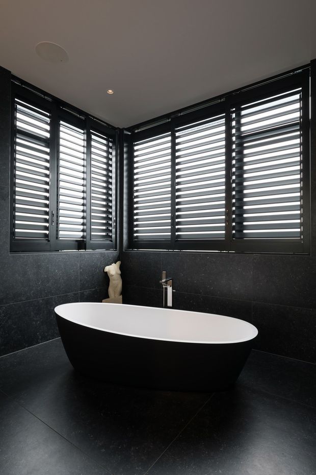 A dramatic contemporary bathroom featuring a freestanding black-and-white bathtub, dark stone-look wall and floor surfaces, a minimalist chrome tap, and corner windows fitted with black horizontal plantation-style shutters. Recessed ceiling lighting and strong monochrome contrast create a luxurious, moody atmosphere.