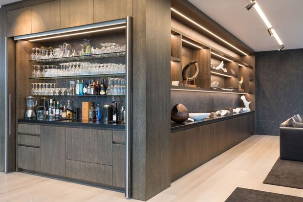 A refined contemporary home bar and display joinery area, featuring dark timber cabinetry, illuminated glass shelving stocked with glassware and spirits, and adjacent recessed display niches with decorative objects. Pale timber flooring, integrated linear lighting, and dark textured wall finishes create a warm, luxurious atmosphere. A partial sofa and rug are visible at the edge of the adjoining living area.