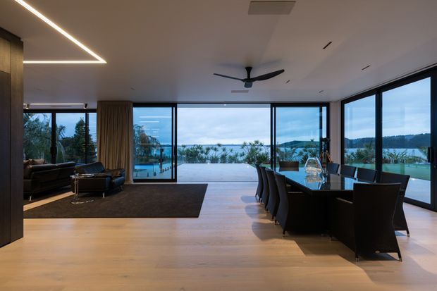 Contemporary open-plan living and dining area with warm light timber flooring, a dark lounge zone, long dining table, sculptural linear ceiling lighting, and extensive floor-to-ceiling glazing opening to a pool terrace and expansive water views.