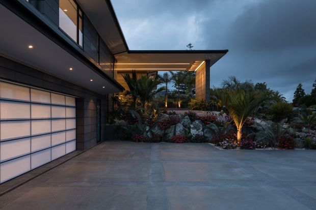Twilight exterior view of a contemporary multi-level residence, showing a broad concrete driveway, integrated garage with translucent segmented door, dark cladding, extensive glazing, projecting roof forms, recessed lighting, and a landscaped entry garden with palms and planted rock beds.
