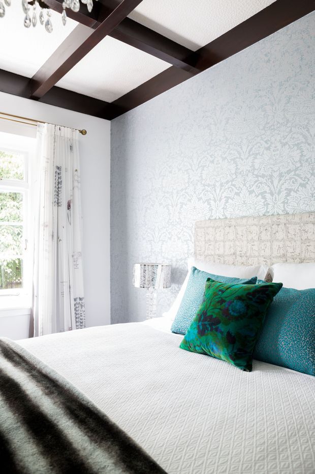 Bright, elegant bedroom with a white quilted bed, upholstered headboard, turquoise and teal patterned cushions, pale blue damask-patterned wallpaper, and white curtains beside a window. Dark stained exposed ceiling beams create a strong architectural contrast, with a crystal chandelier and mirrored bedside lamp adding refined decorative details.