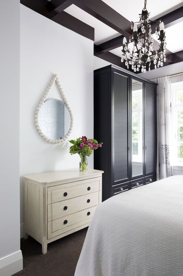 Bright, elegant bedroom with a white upholstered bed, dark carpet, a distressed ivory chest of drawers, tall black built-in wardrobes, and a large ornate crystal chandelier. White walls and curtains contrast with exposed dark ceiling beams, while a decorative shell-framed mirror and flowers add softness.