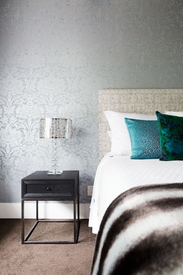 A styled bedroom corner featuring a bed with a tall textured upholstered headboard, crisp white bedding, vivid teal and blue accent cushions, and a dark wood bedside table with a clear glass lamp. The wall has ornate pale grey patterned wallpaper, and the room is finished with neutral carpeting and a dark-and-white throw.
