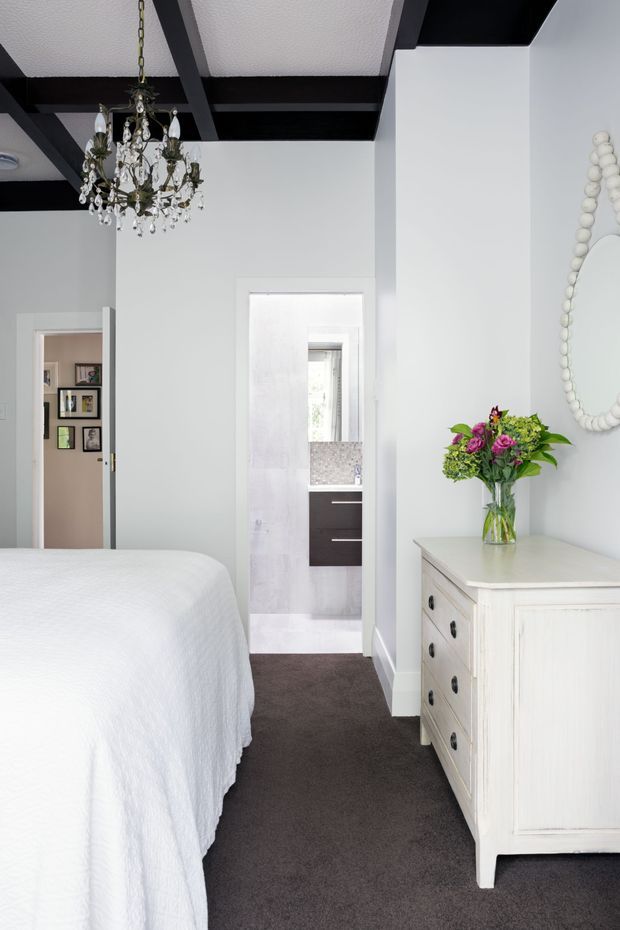 Bright traditional bedroom with a partially visible bed, white walls, dark charcoal carpet, exposed dark ceiling beams, and an ornate crystal chandelier. A distressed white dresser with black hardware sits beneath a beaded oval mirror, while an open doorway reveals a bathroom with a vanity and tiled surfaces.