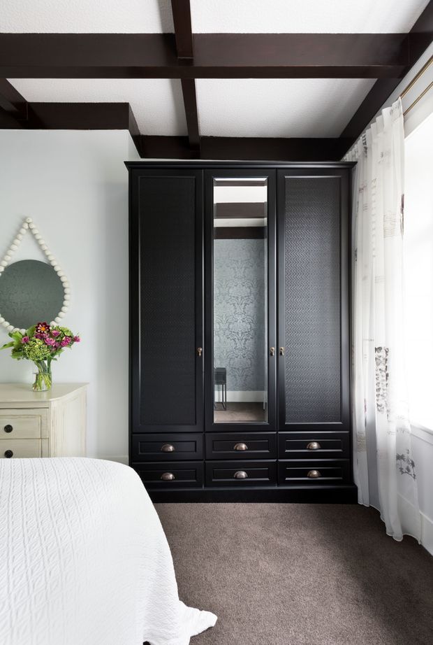 A traditional bedroom corner featuring a large built-in black wardrobe with mirrored central door, textured panelled doors, and multiple drawers. Dark exposed ceiling beams contrast with pale walls and a soft carpeted floor. A partially visible white bed, cream dresser, decorative mirror, floral arrangement, and sheer curtains complete the room.