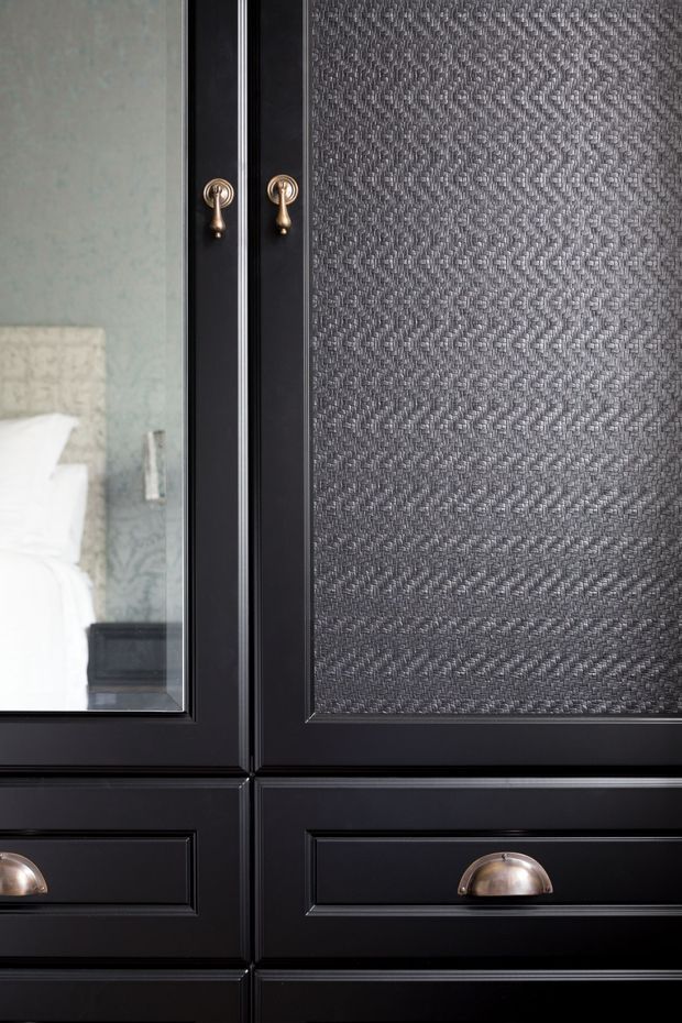 Close-up of a dark black built-in wardrobe or cabinet with raised panel detailing, brushed brass drop handles, and brass cup pulls. One upper panel is glazed and reflects a bedroom with white bedding, while the adjacent panel contains textured dark grey fabric or woven inlay. The design is formal, tailored, and high-contrast.