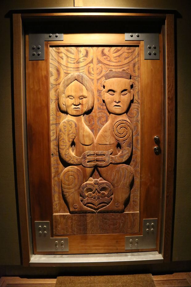 A close-up interior view of an ornate carved timber door featuring a detailed Māori-inspired figure and swirling whakairo-style patterns. The door is framed in dark wood with prominent metal corner plates, hinges, and hardware, set within a dimly lit entry or passage area.