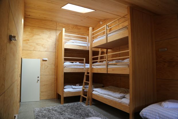 Compact bunk-bedroom interior with multiple built-in timber bunk beds, a central ladder, warm plywood wall and ceiling lining, a recessed rectangular ceiling light, and grey carpeted flooring. The room is functional and densely arranged for shared sleeping accommodation.
