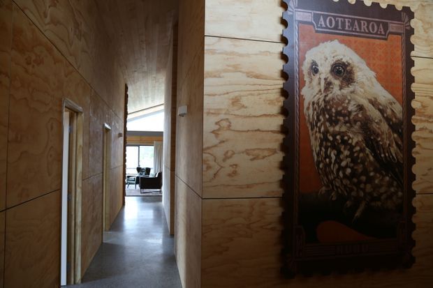 A narrow residential hallway lined with warm-toned plywood wall panelling and matching timber door frames. The corridor leads toward a bright living area with glazing and seating visible at the far end. A large framed New Zealand-themed owl artwork forms a feature on the right wall, while the floor has a smooth dark grey finish.